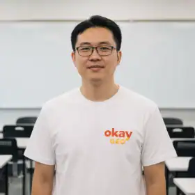 A young man with glasses wearing a white OkayGEO t-shirt, standing in a modern classroom or office setting with black chairs behind him.